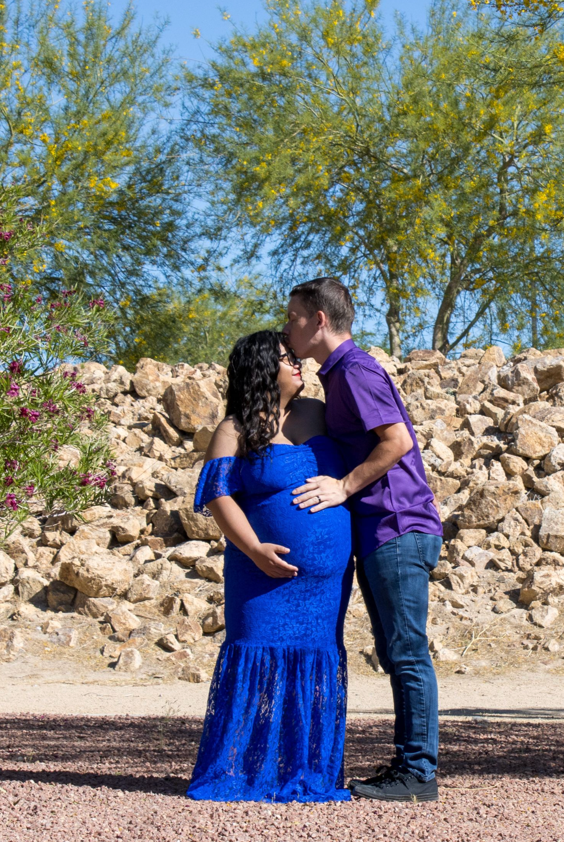 Pregnant woman in blue lace gown kissed by partner in purple shirt outdoors with rocks and green trees.