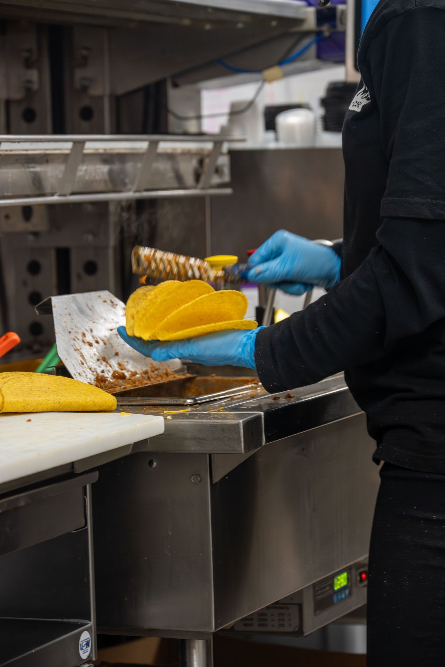 Taco Bell worker in blue gloves filling taco shells with seasoned meat at the kitchen station.