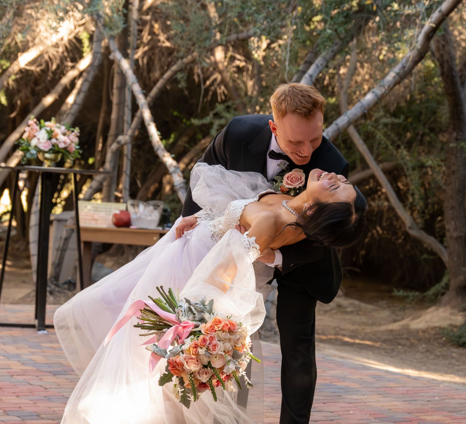 Groom dips bride in elegant wedding attire outdoors, bride holding colorful bouquet near rustic teepee backdrop.