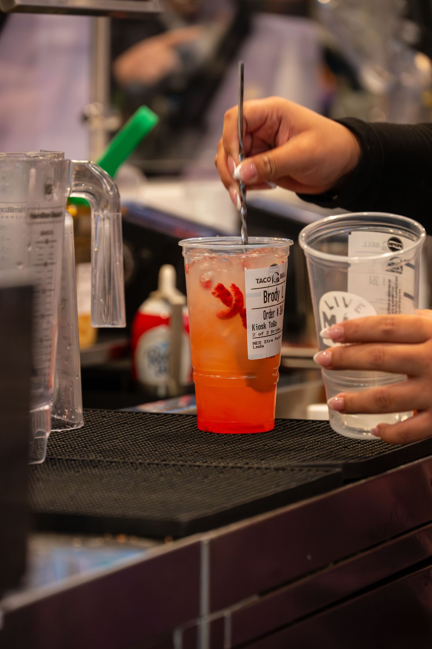 Bartender stirring a vibrant red gradient cocktail in a plastic cup at a busy bar counter.
