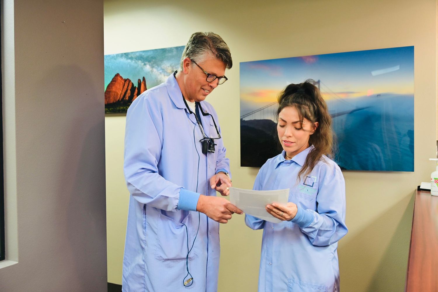 Male dentist and female dental assistant review treatment documents together in clinic corridor.