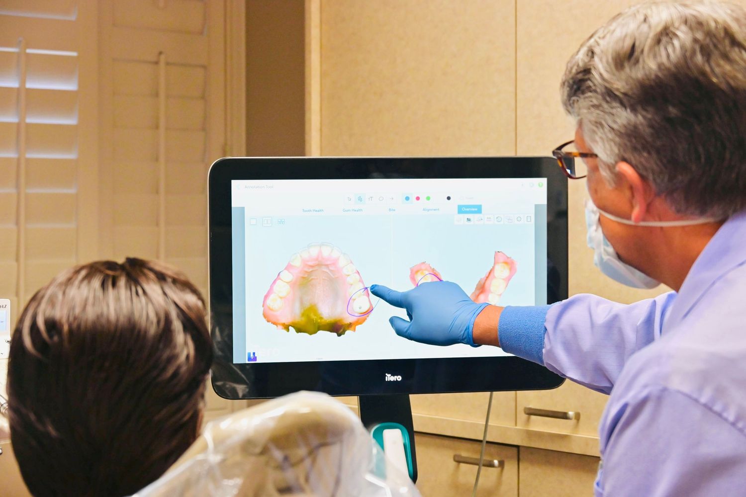 Dentist in blue gloves showing a patient a 3D dental scan on a monitor during a consultation.