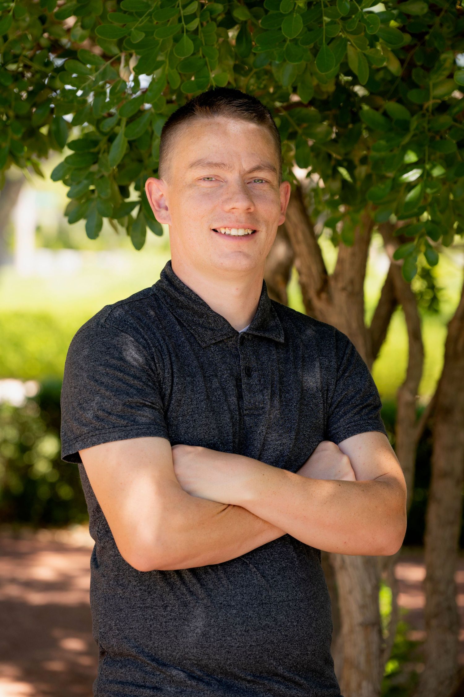 Smiling young man in dark polo shirt standing with arms crossed under green leafy tree outdoors.