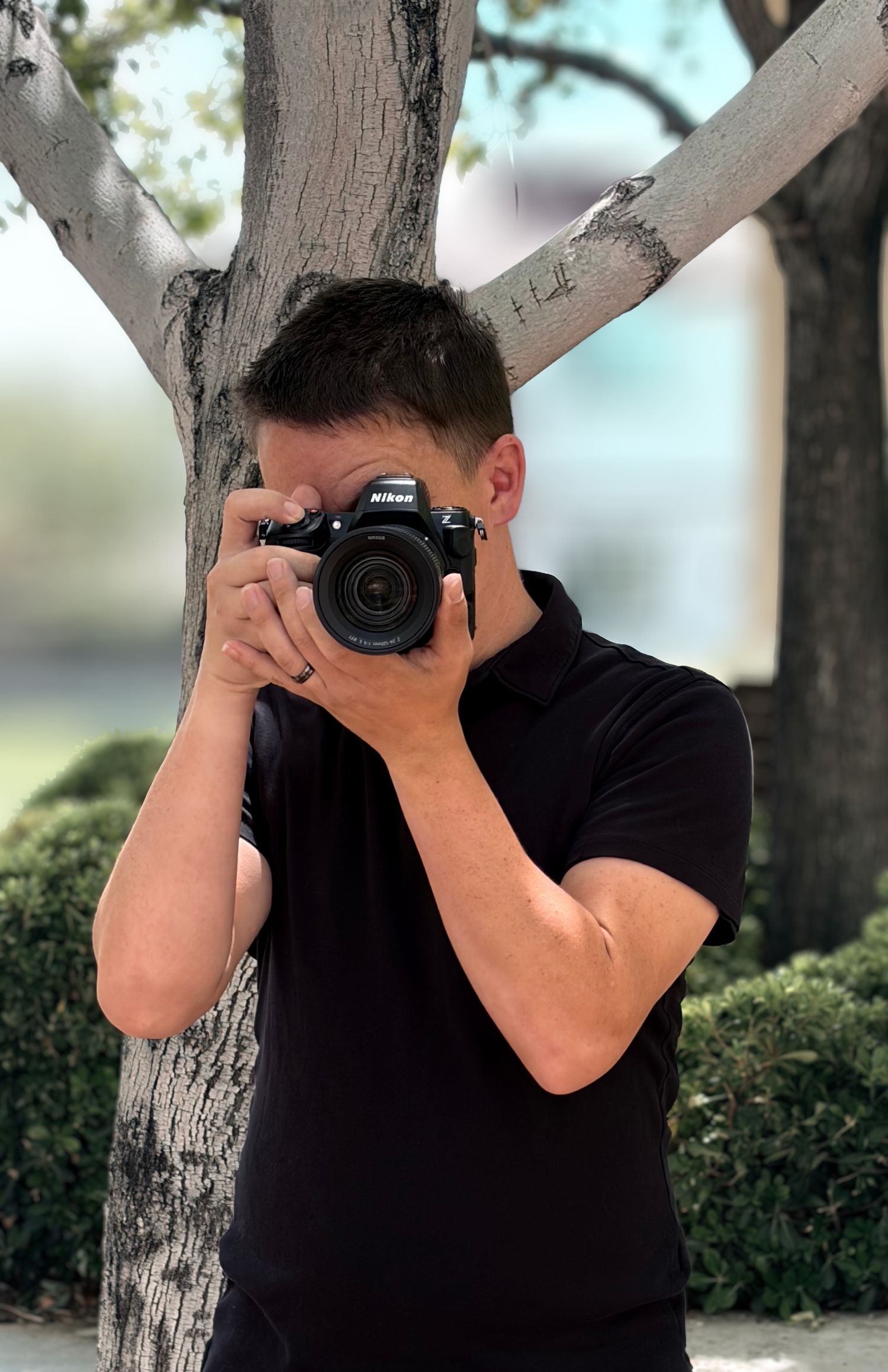 Male photographer in black polo aiming Nikon camera at viewer while standing beside a tree trunk.