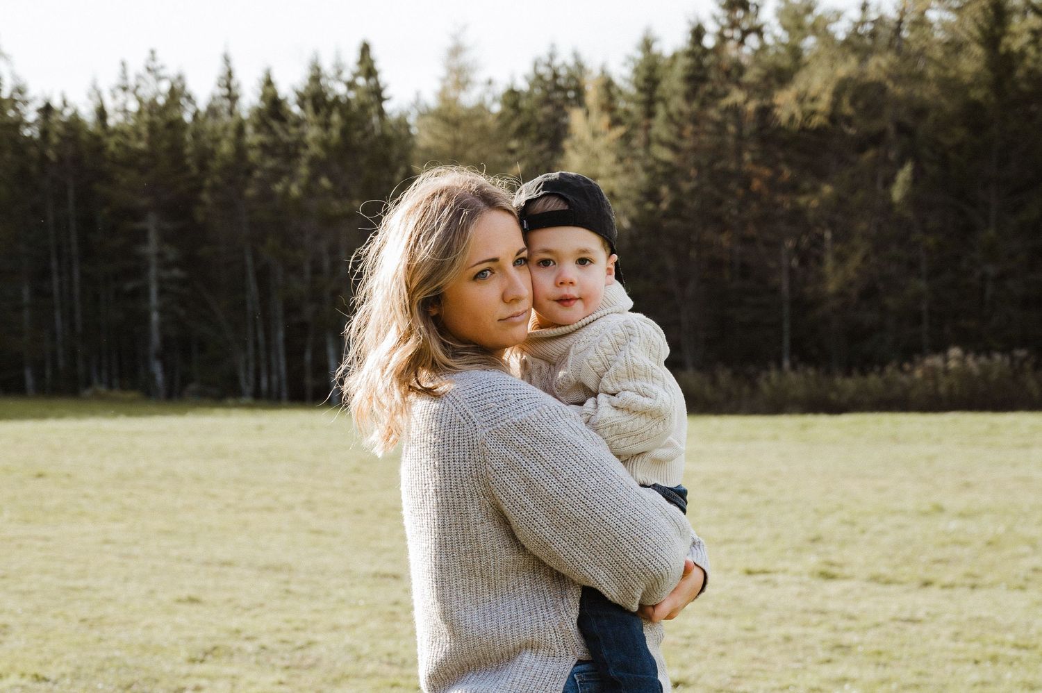Person holds small child while standing in a sunny field with pine trees in background.