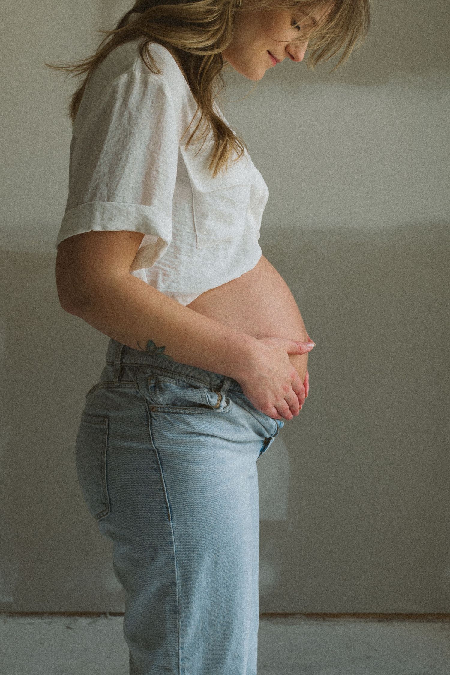 Side view of an expectant mother in a white shirt and jeans gently cradling her pregnant belly in soft lighting.