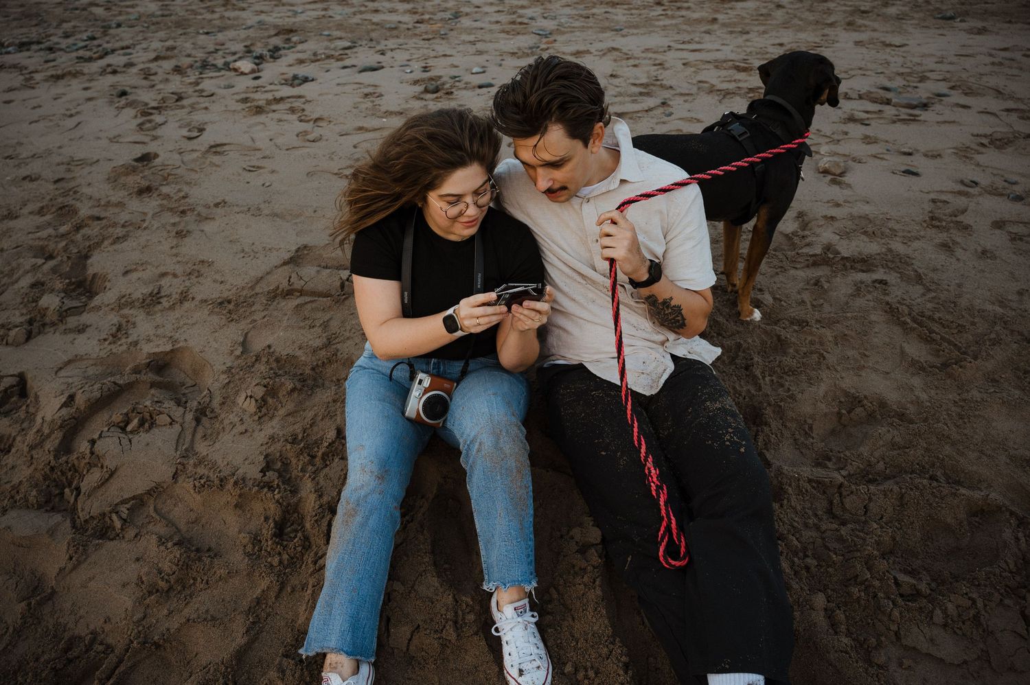 Two people sitting on sandy beach looking at photos while their dog stands nearby on leash.