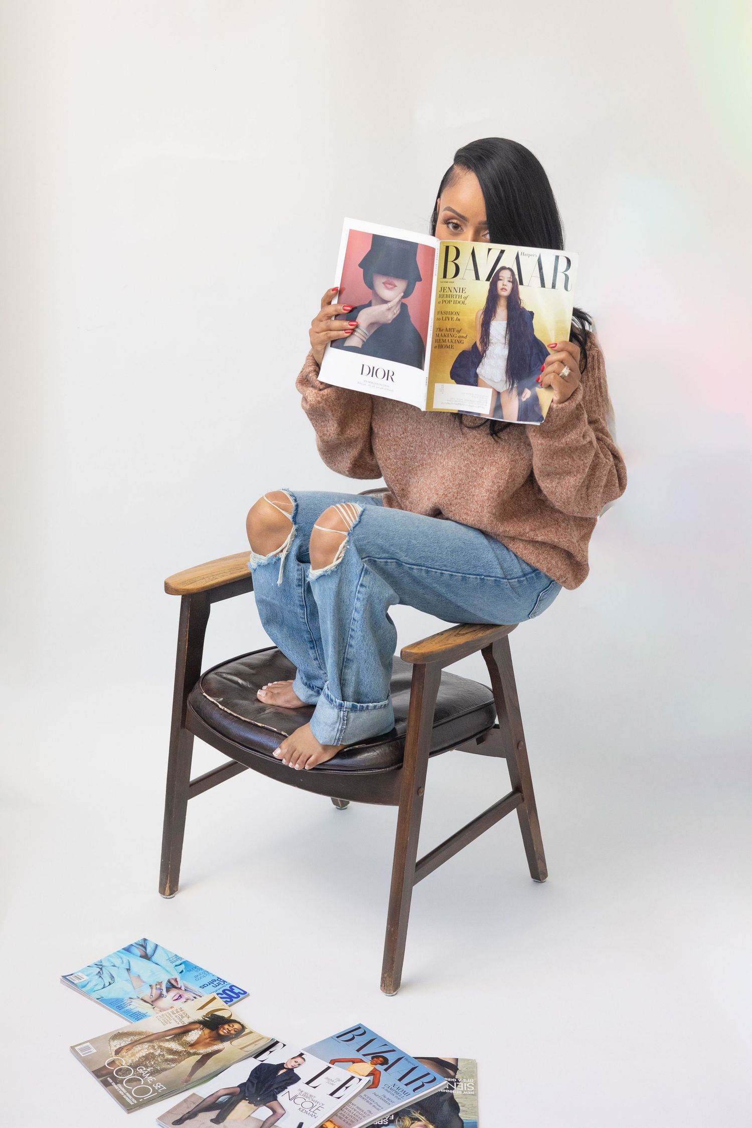 Woman sitting in wooden chair reading Harper's Bazaar magazine, wearing ripped jeans and brown sweater, magazines on floor.