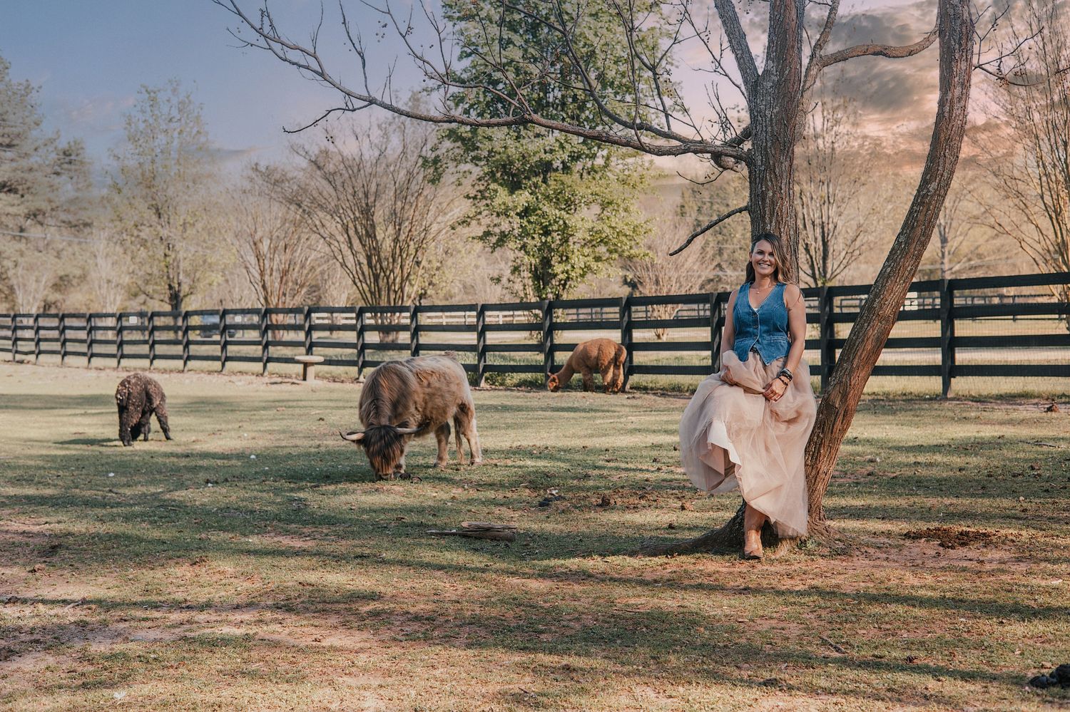 Woman in skirt leans against tree near grazing sheep on a sunny farm.
