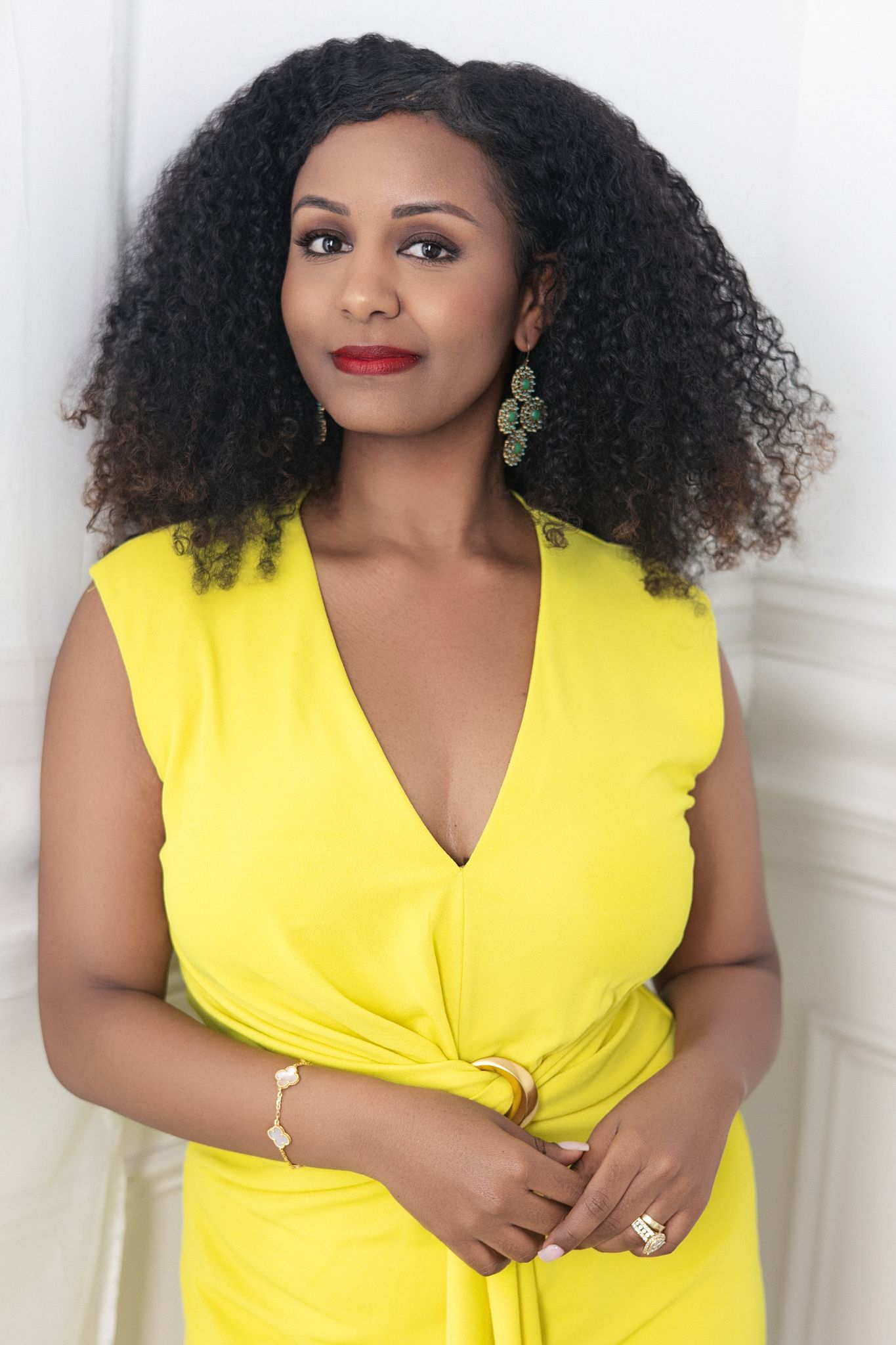 Woman in yellow dress with natural curly hair, red lips, and gold jewelry poses confidently against white wall.