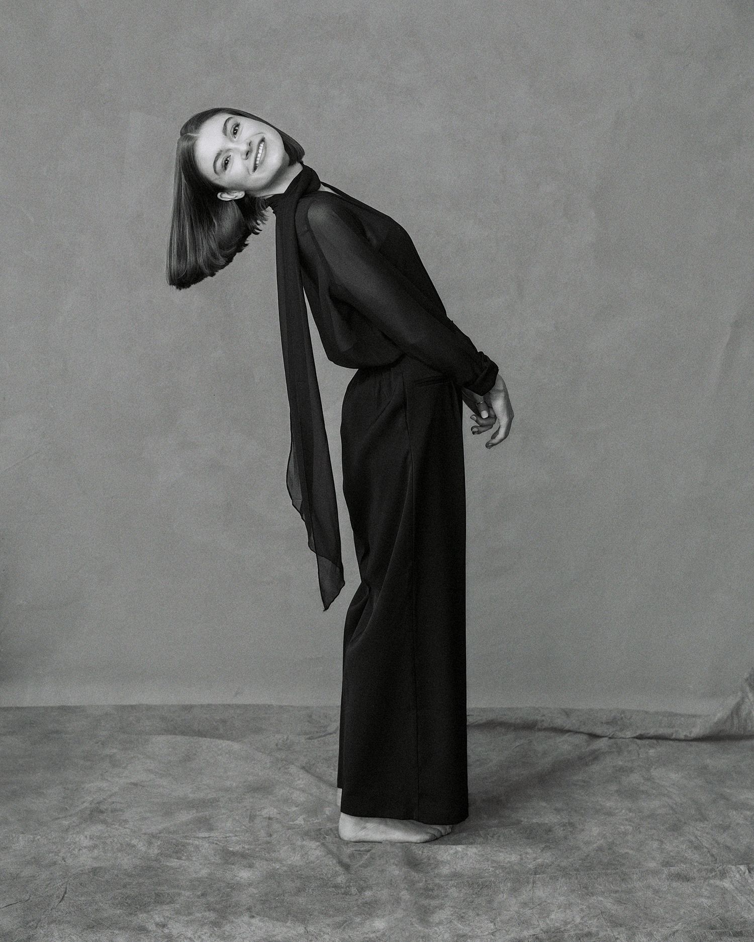 Black and white photo of a woman in elegant black wide-leg jumpsuit with scarf, posing gracefully in studio.