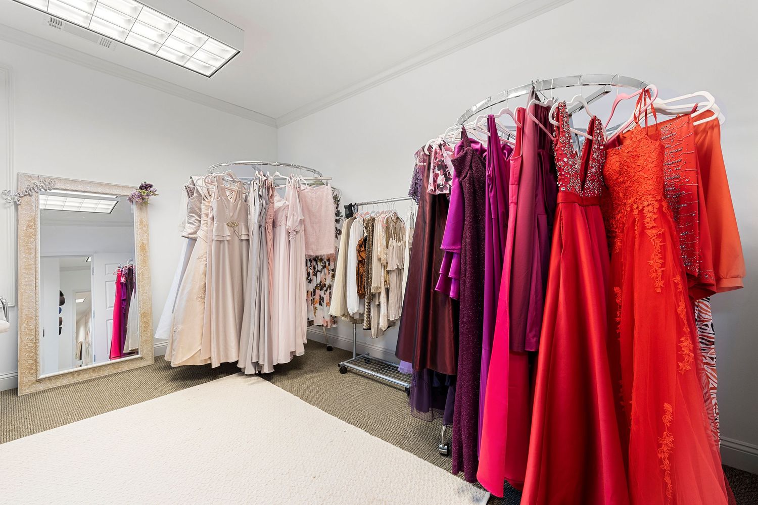 Boutique dressing room with racks of formal gowns in pink, purple, and red hues with mirrors.