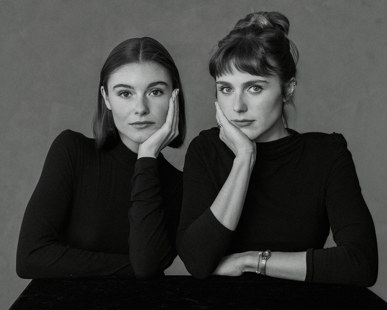 Black and white portrait of two young women in black turtlenecks, resting chins on hands, studio background.