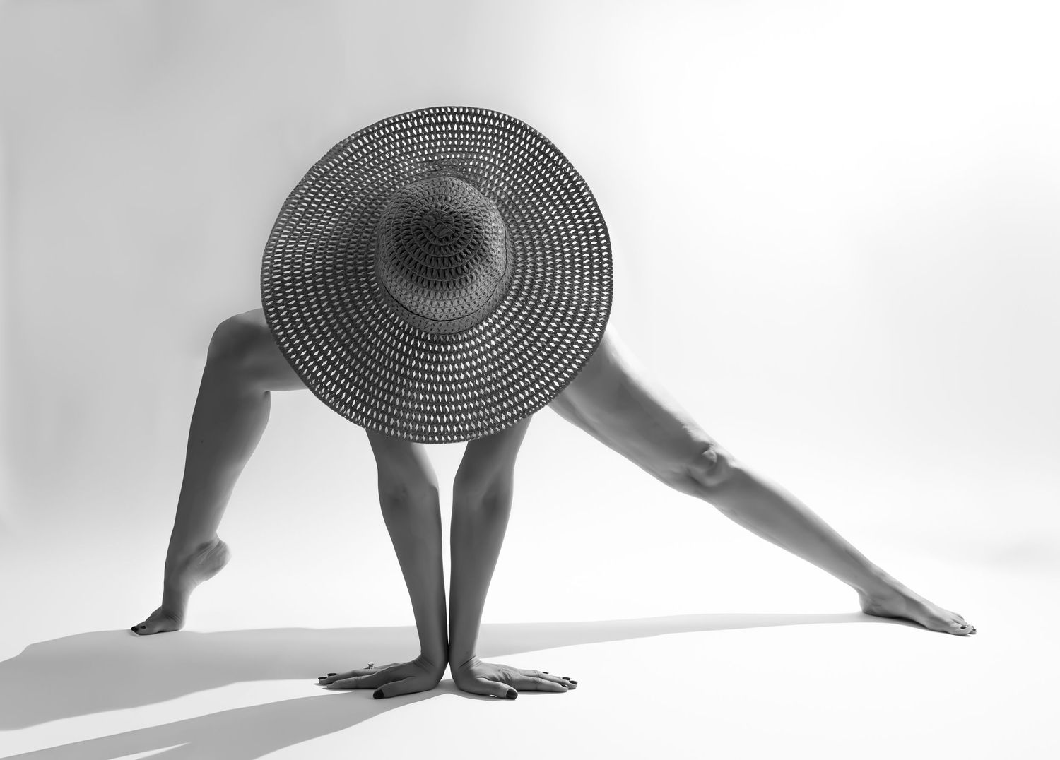 Black and white photo of a person in a wide-brim straw hat doing a yoga pose, bending forward gracefully.