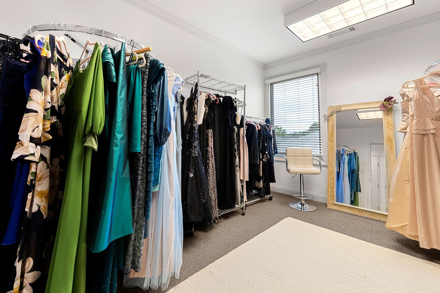 Dressing room with racks of elegant gowns in green and dark tones, mirror, and styling chair.