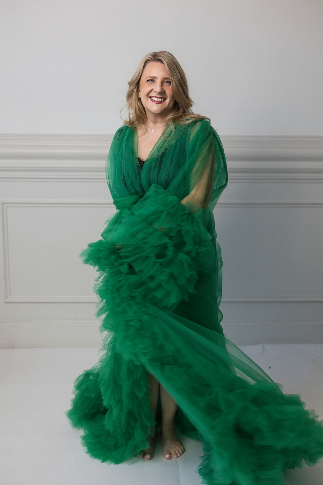 Smiling blonde woman posing in a flowing emerald green tulle gown against a white studio backdrop.