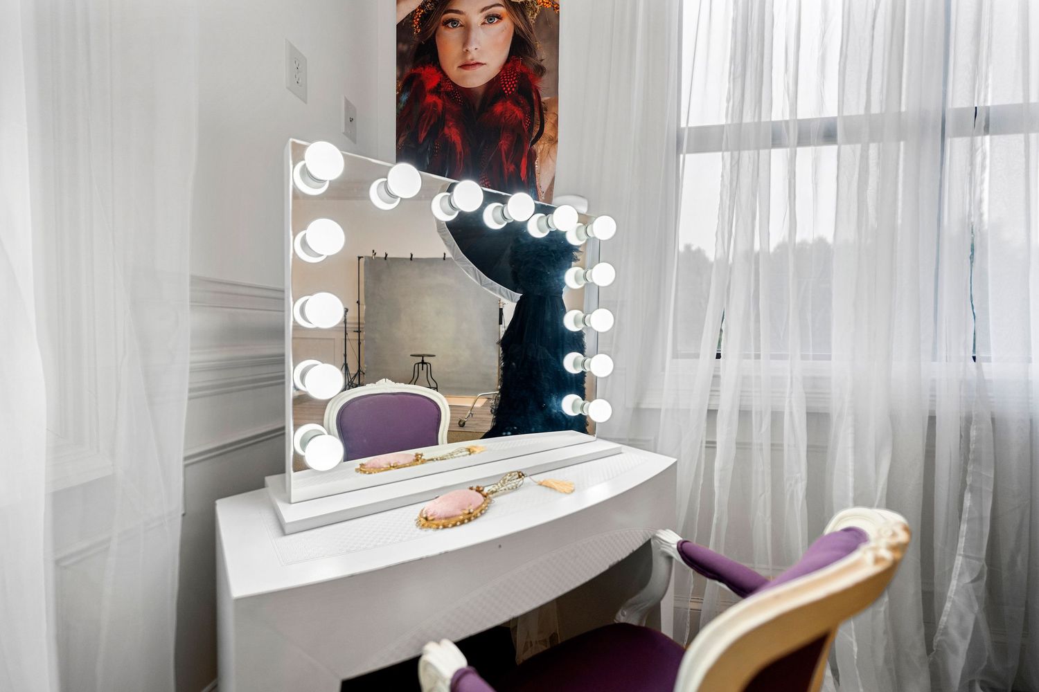 Hollywood vanity makeup mirror with bulb lights on white desk beside sheer curtains.