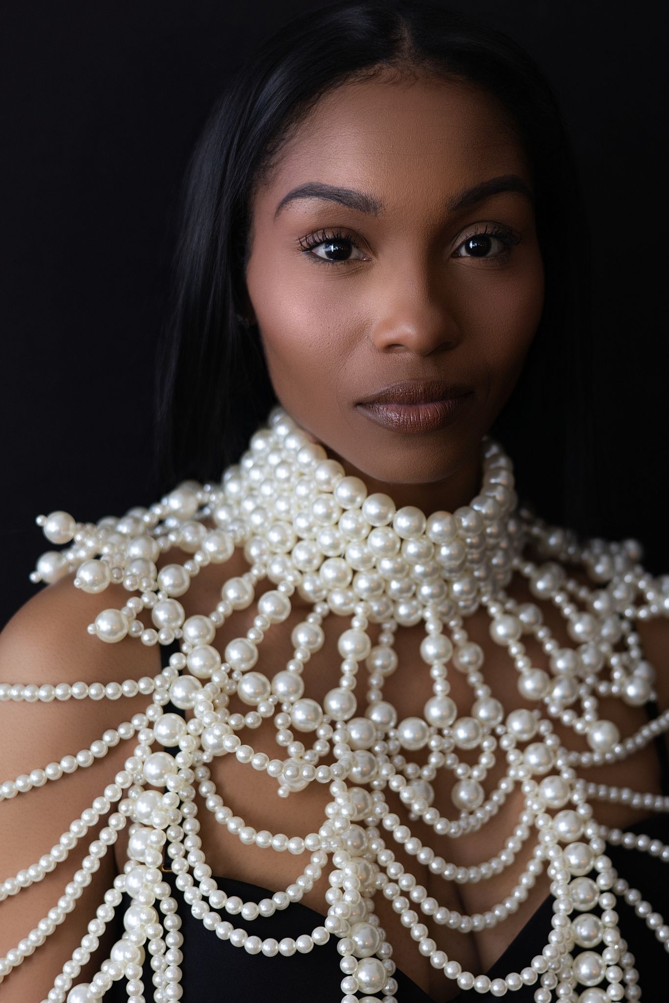 A young Black woman wearing an elaborate pearl necklace draping over her shoulders, posing against a dark background.