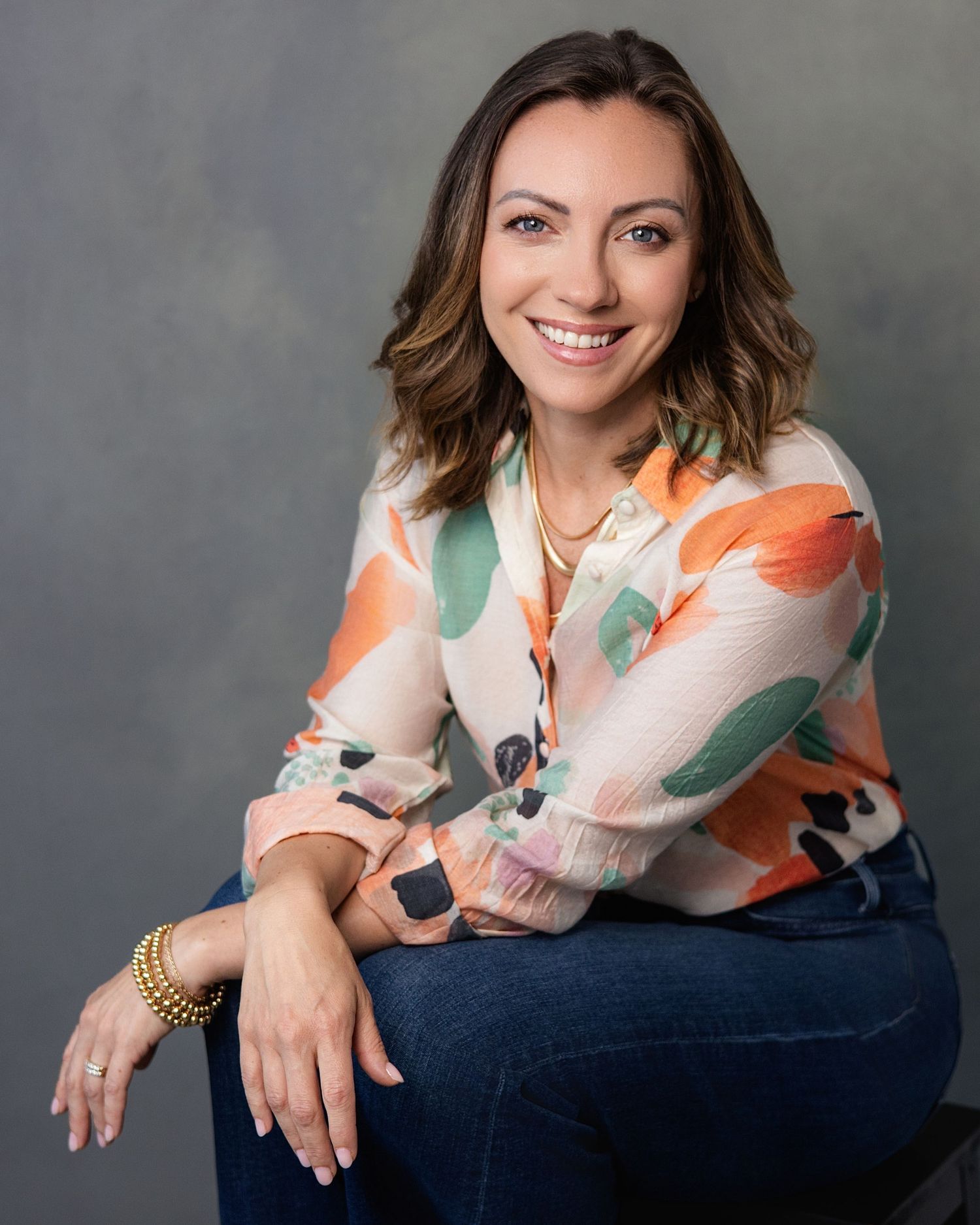 Professional woman smiling in floral blouse and jeans seated for studio headshot.