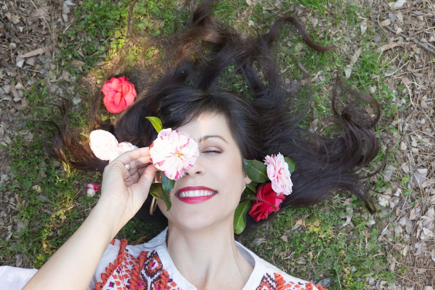 Woman lying on grass with dark hair spread out, holding pink flowers over her eye, smiling joyfully.