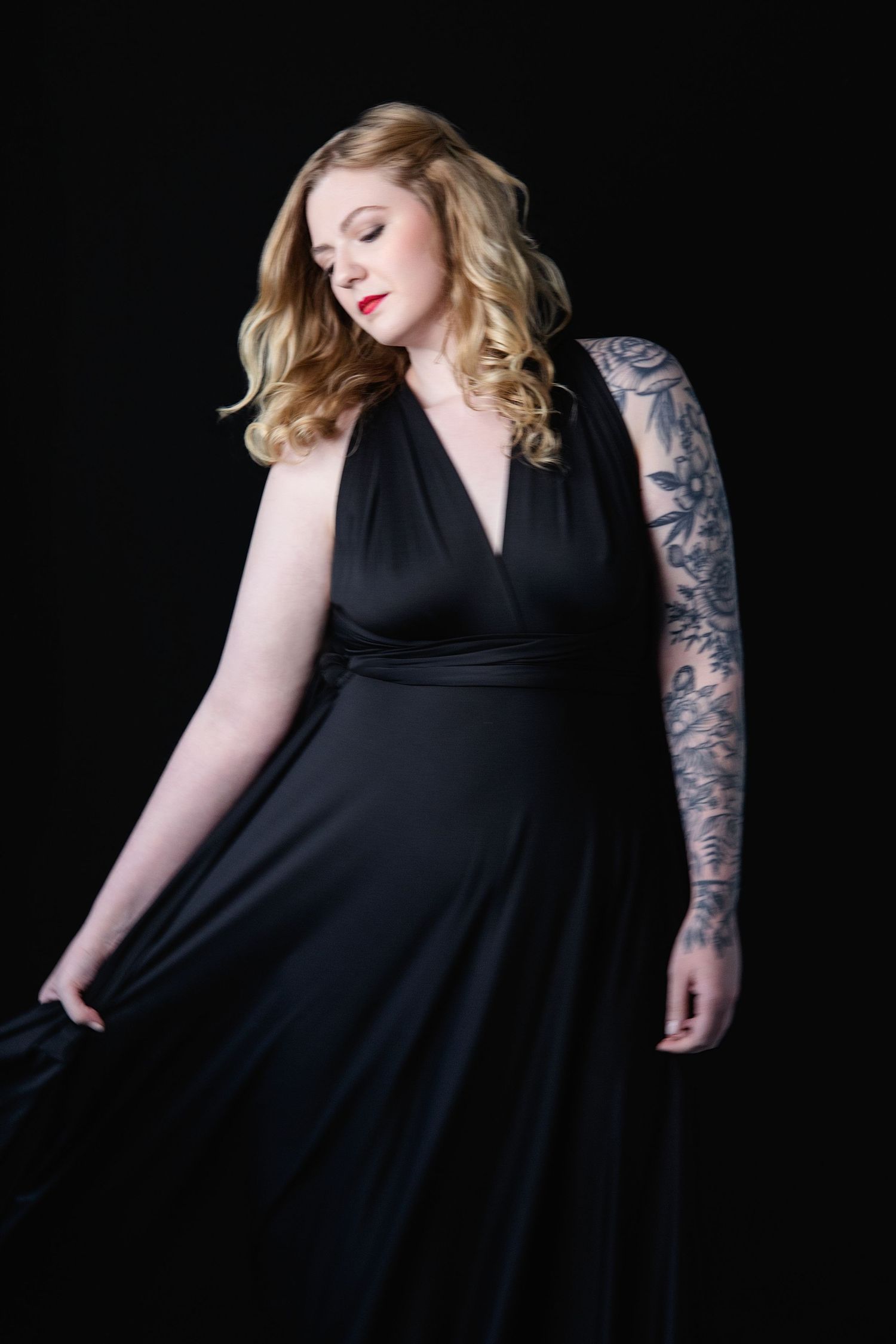 Plus-size woman in black wrap dress with sleeve tattoos poses elegantly against dark background.