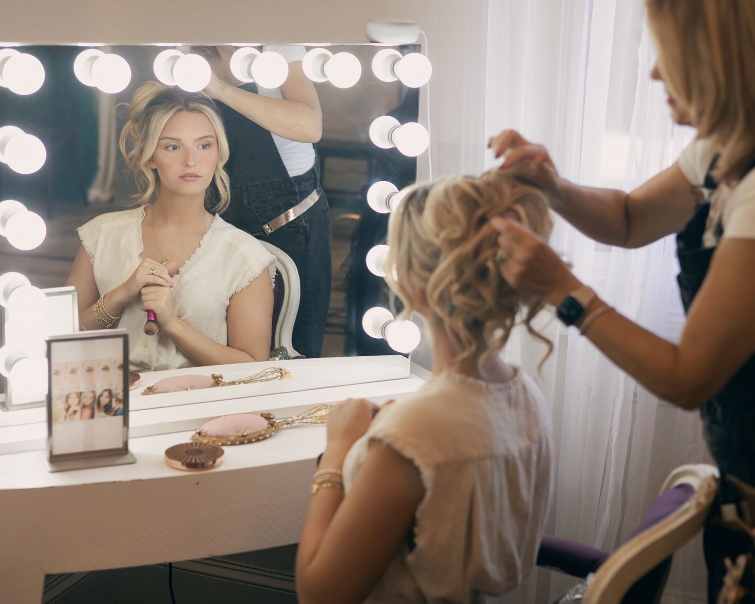A bride sits at a vanity mirror with Hollywood lights while a hairstylist styles her elegant updo in a bright room.