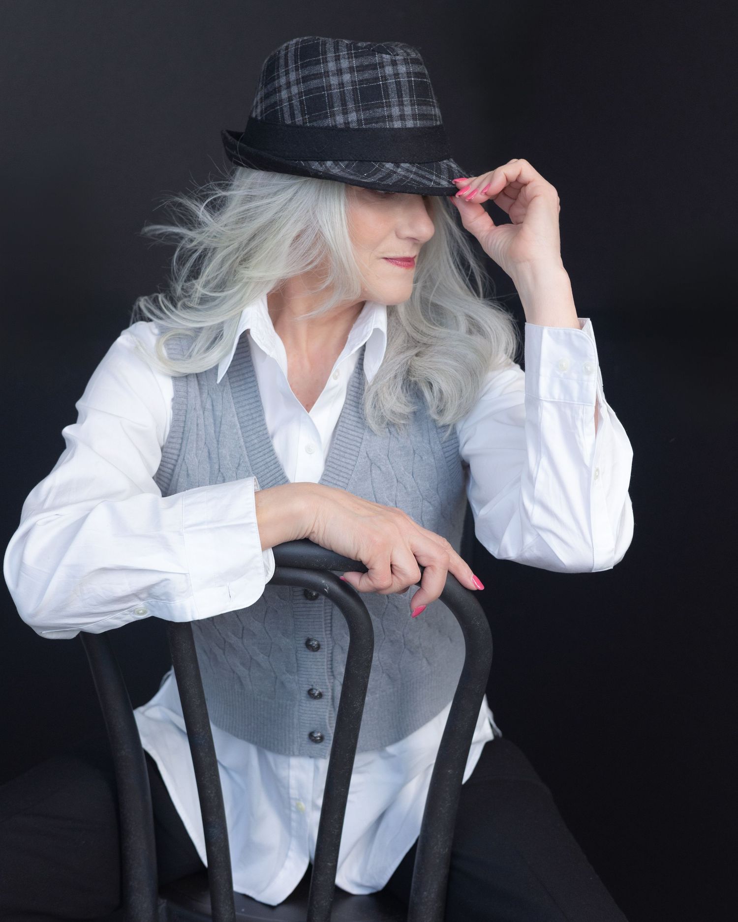 Elegant woman with silver hair wearing a plaid fedora, white blouse, and gray vest, seated on a black chair.