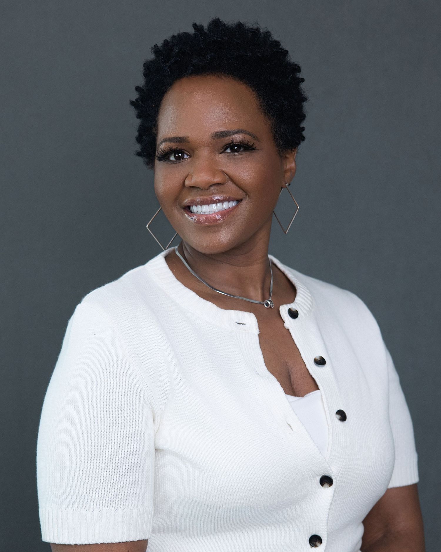 Professional headshot of a smiling woman in a white knit top with geometric earrings against a gray background.