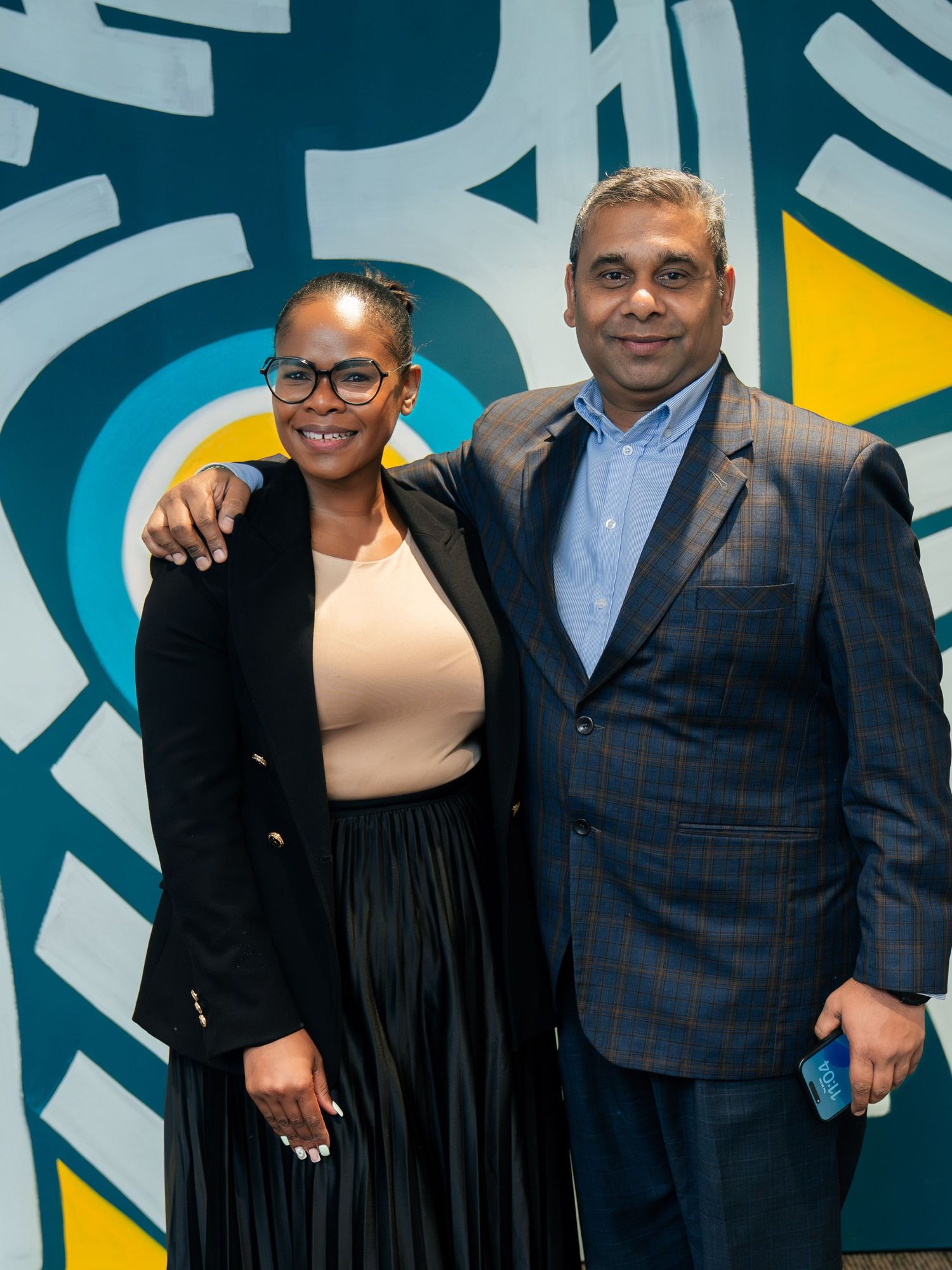 Two professionals smiling together in front of a colorful abstract mural at a corporate event.