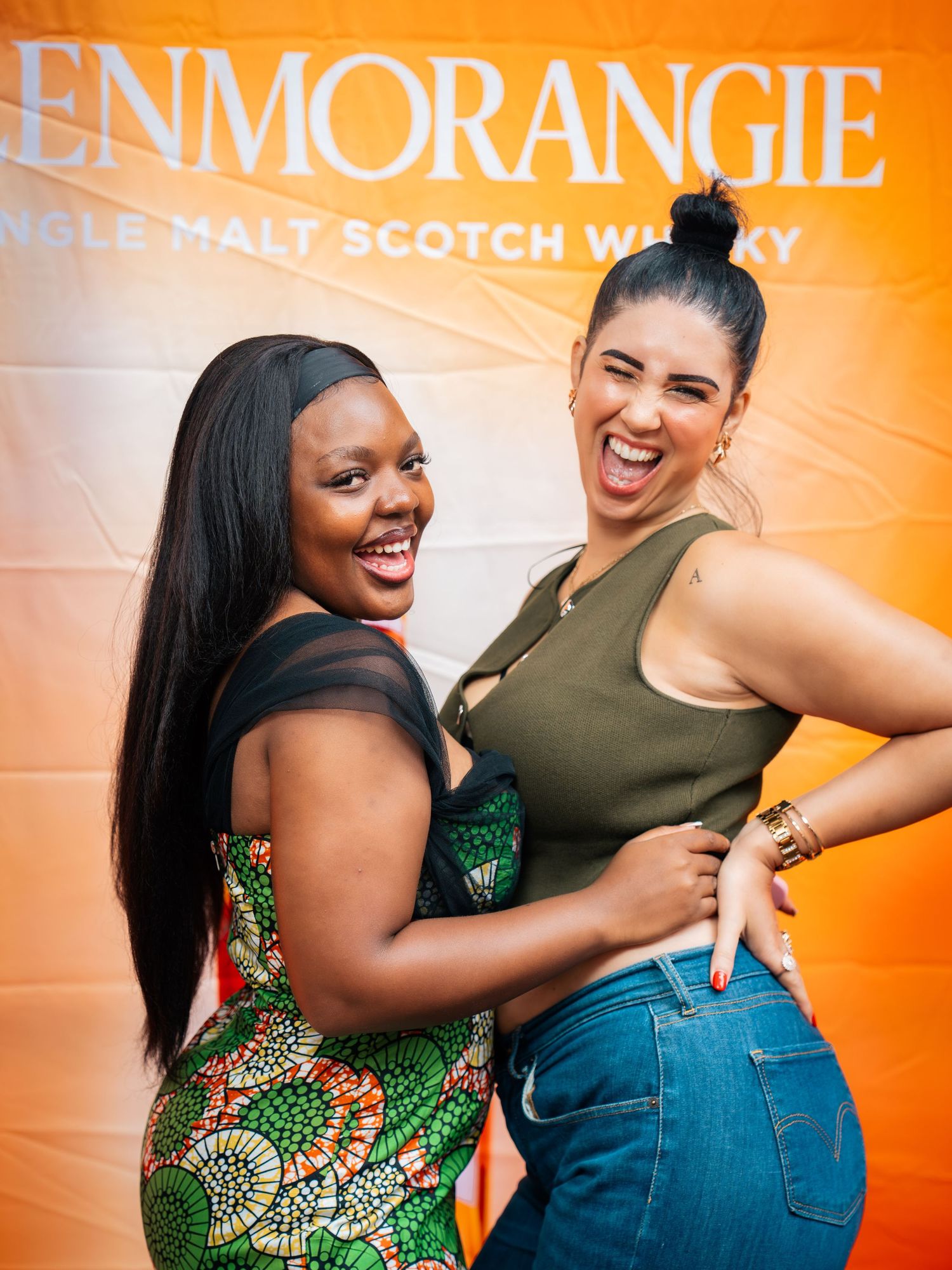 Two women smiling and posing in front of a Glenmorangie Single Malt Scotch Whisky orange banner.