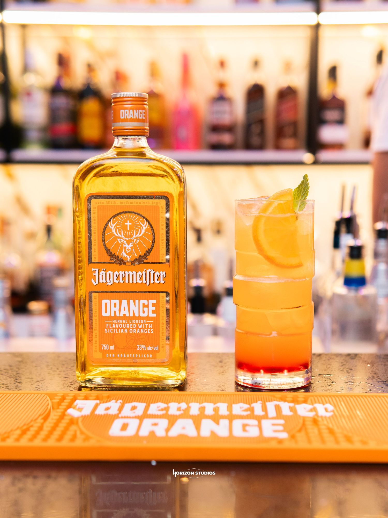 Jagermeister Orange liqueur bottle beside a tall cocktail garnished with orange slice on a branded orange bar mat.