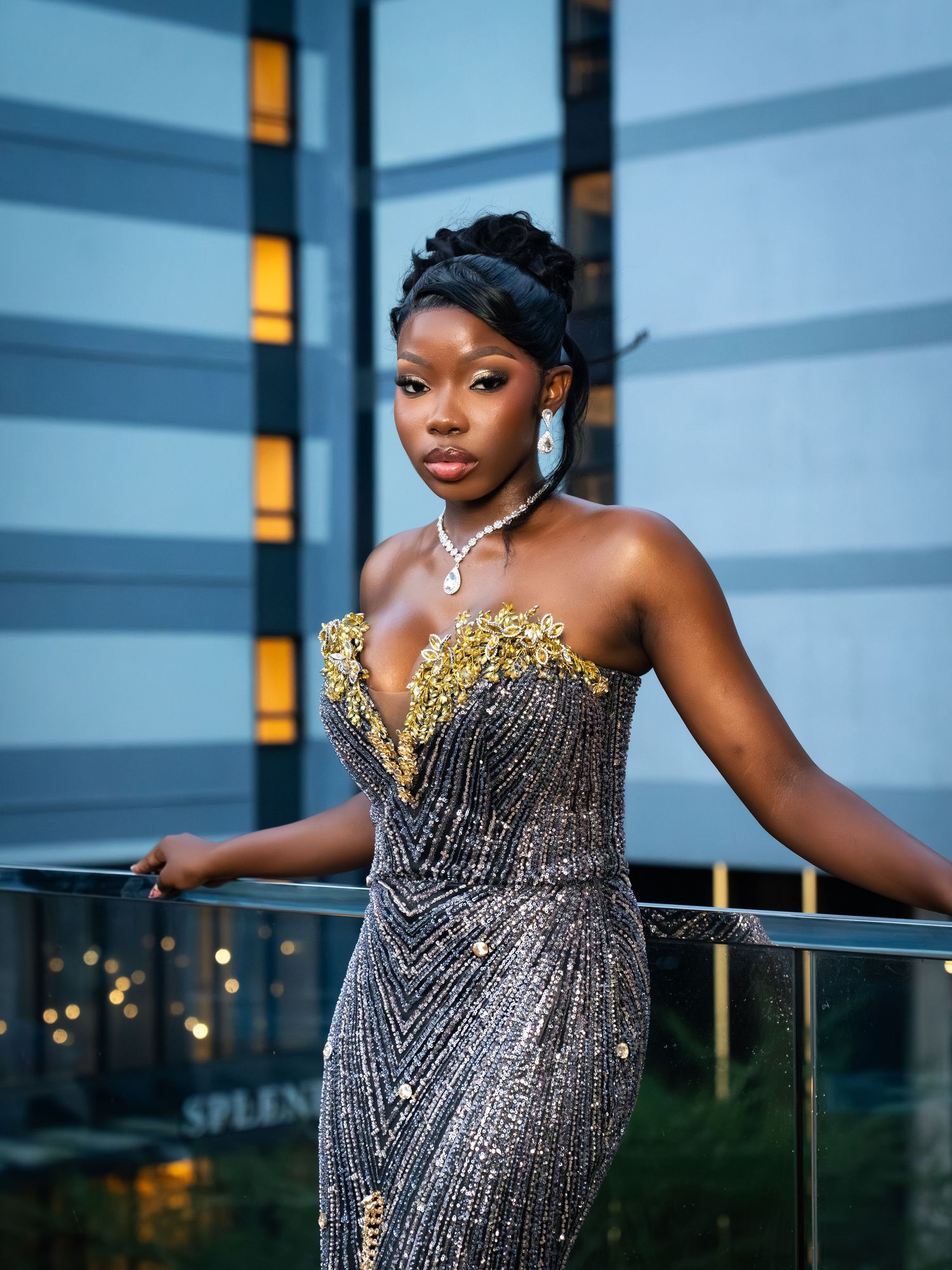 Elegant woman in a sparkling silver and gold strapless gown with pearl necklace posing on a balcony at dusk.