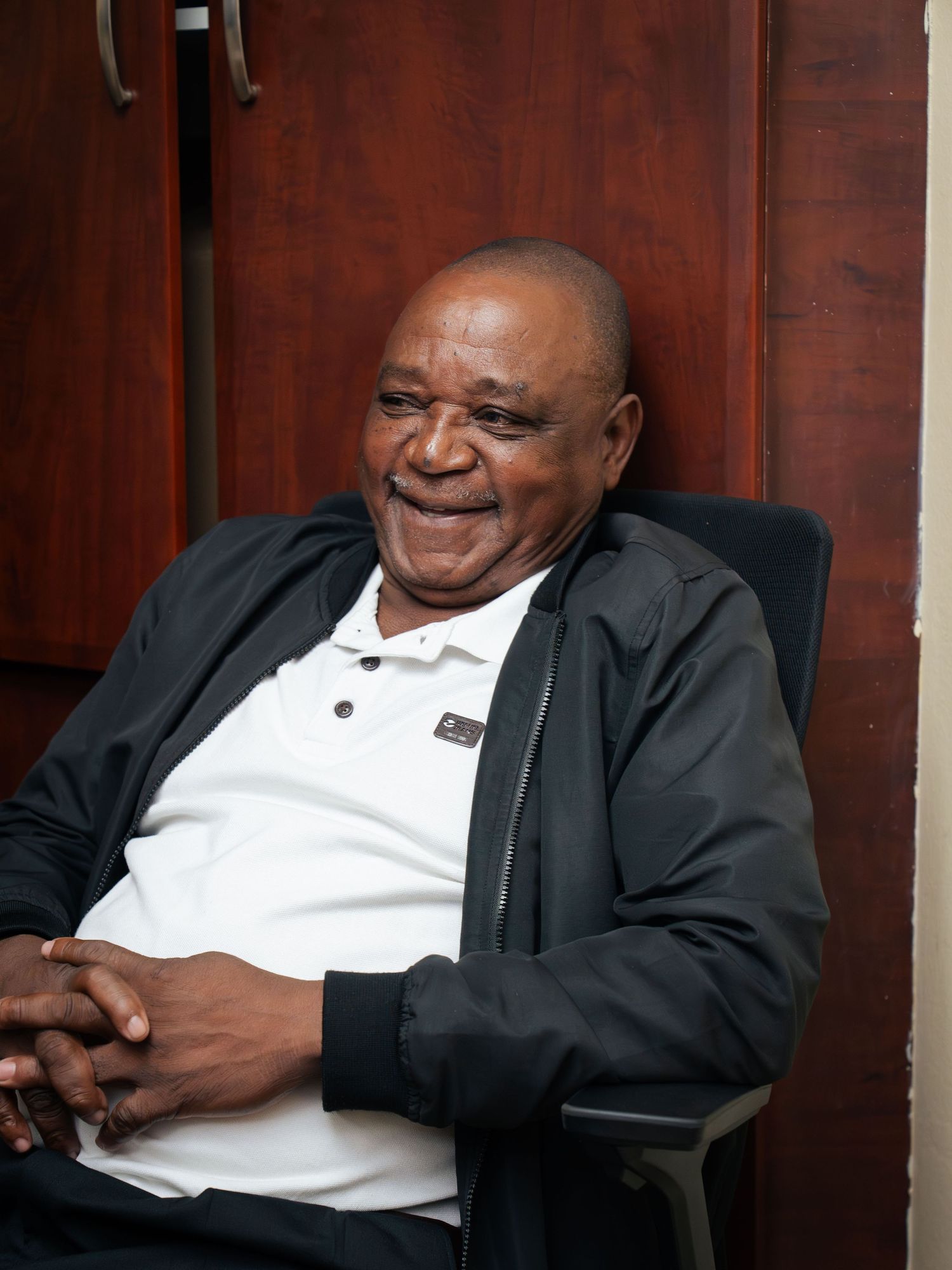 Elderly man smiling warmly while seated in a chair, wearing a black jacket over a white polo shirt.