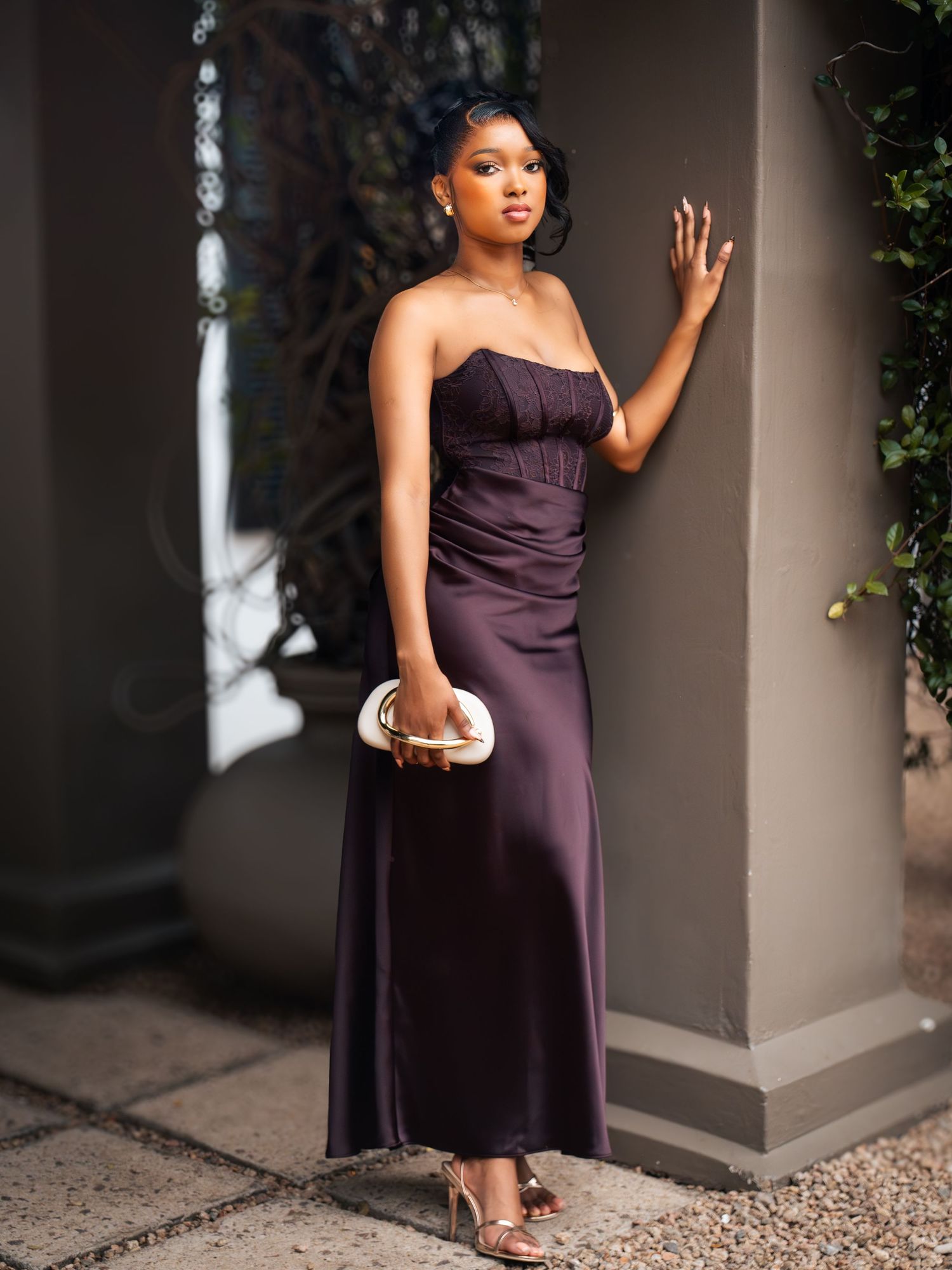 Woman in strapless dark purple satin maxi dress holding clutch, posing elegantly in heels against a column.