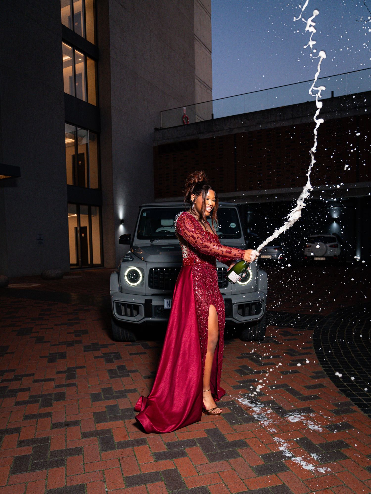 Woman in red gown spraying champagne in front of a Mercedes G-Wagon at night.