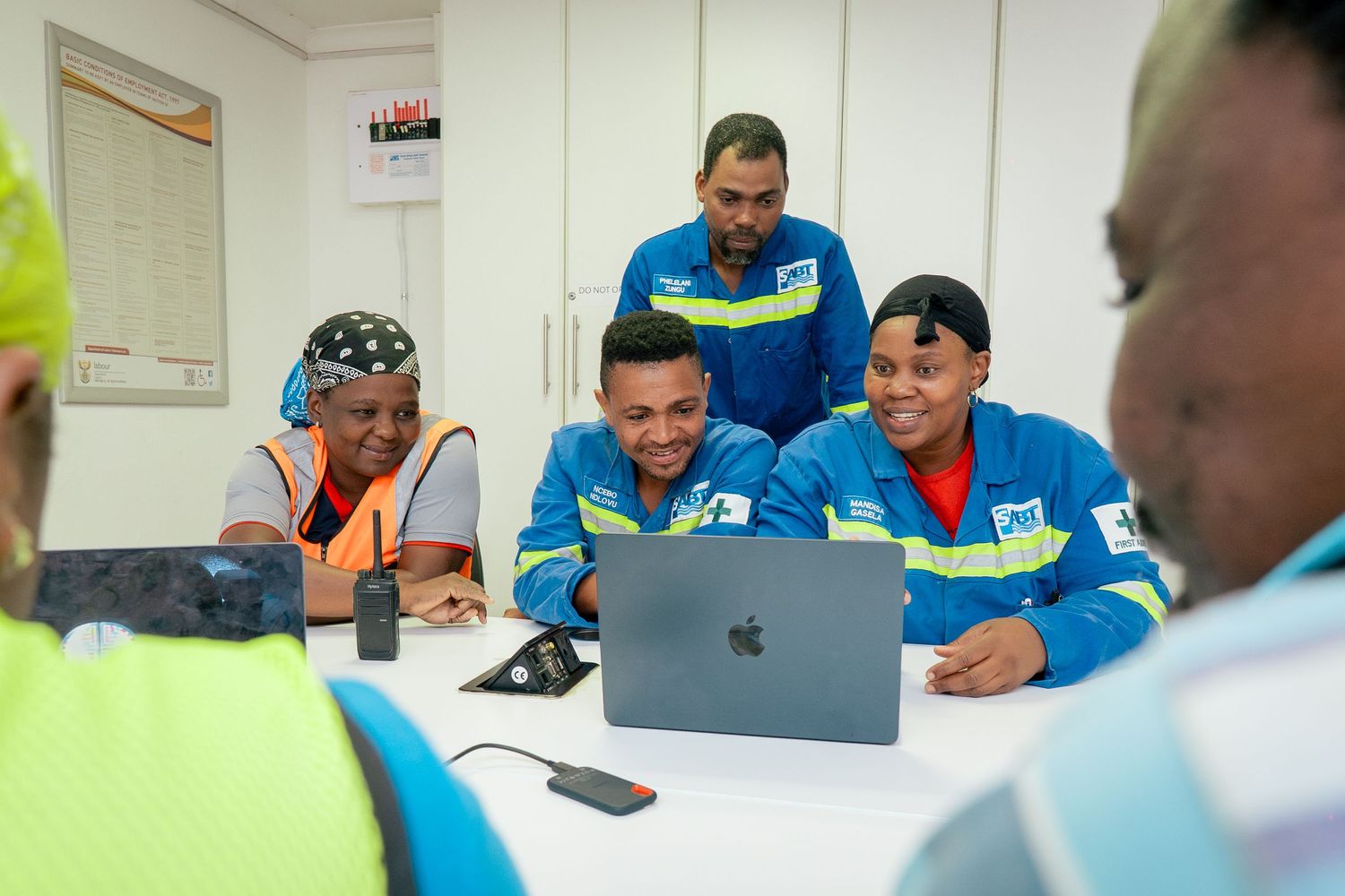 Workers in blue uniforms collaborate around a laptop in an office, smiling and engaged in a team meeting.