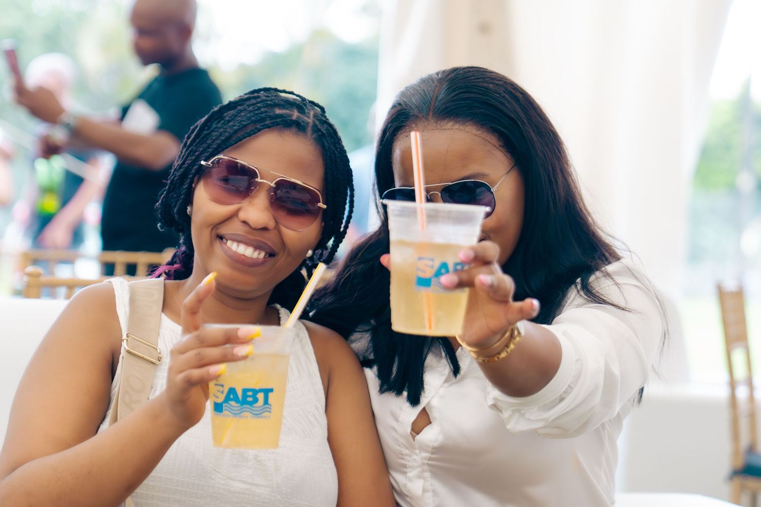 Two smiling women holding ABT branded drinks outdoors at a sunny event.