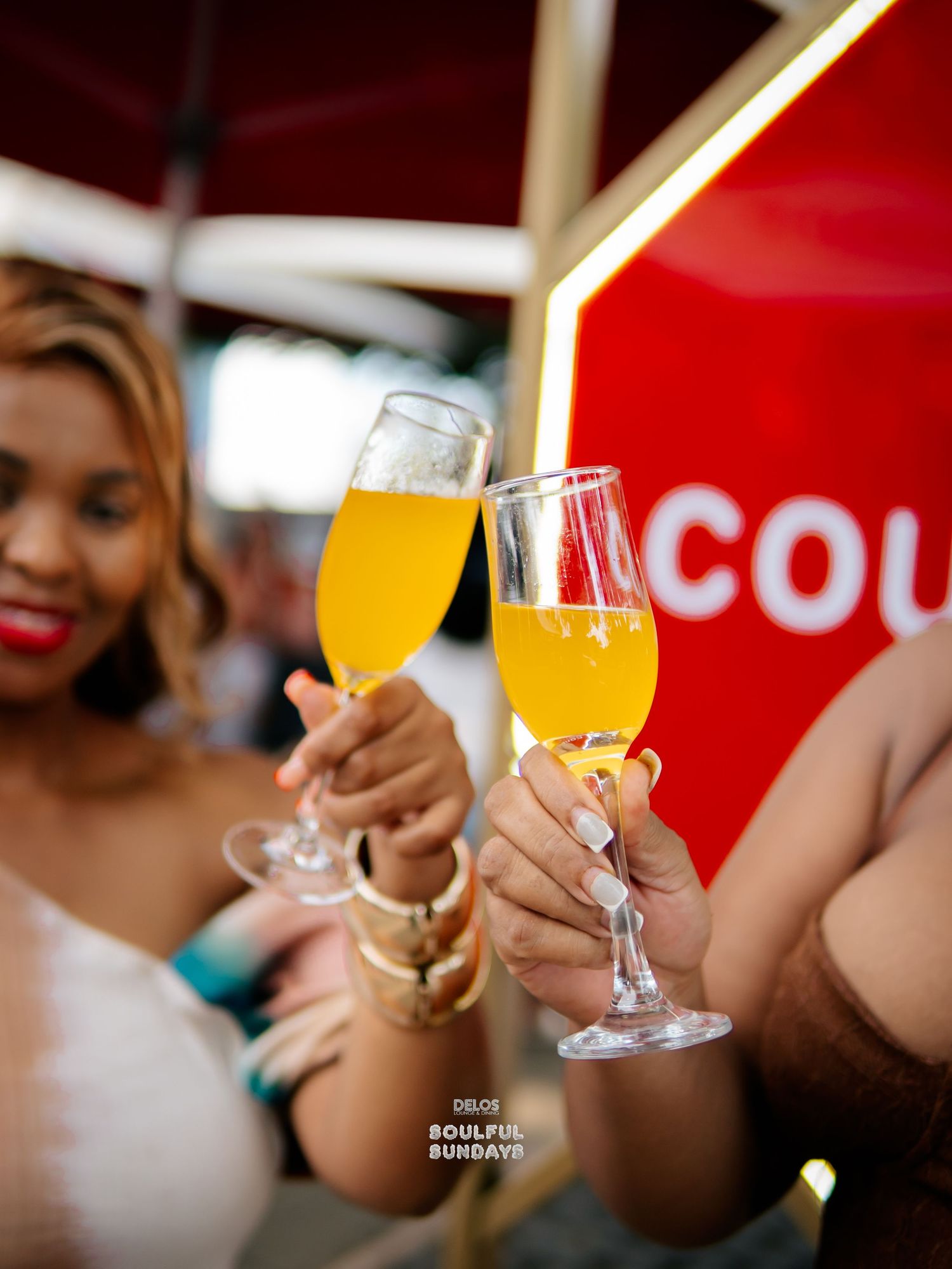 Two women toasting with champagne flutes filled with orange mimosas at a vibrant outdoor brunch event.