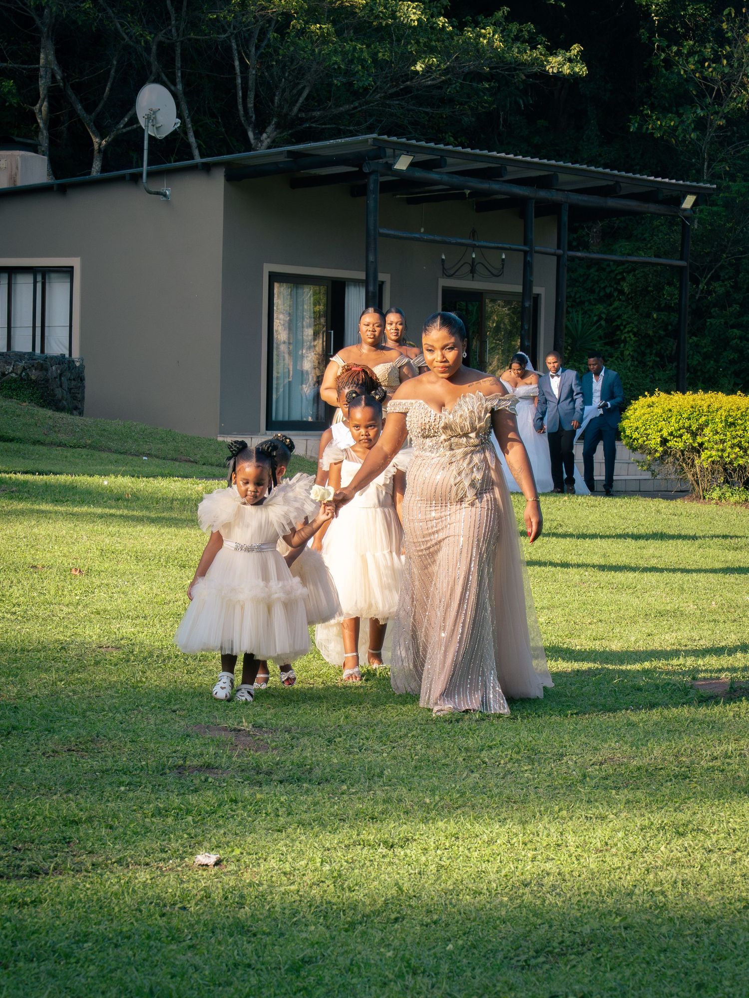 Bride in elegant gown walks with flower girls on green lawn at outdoor wedding ceremony.