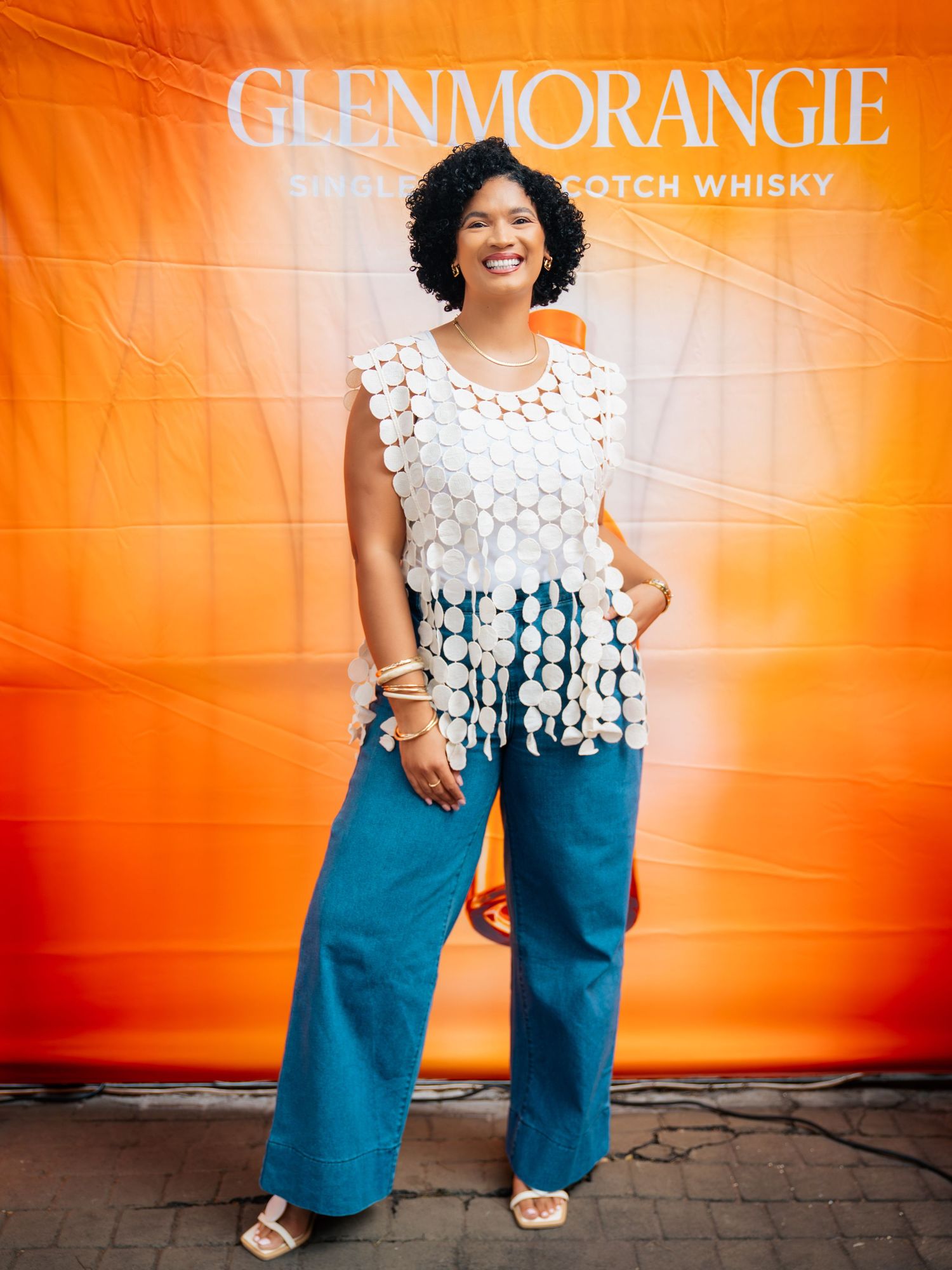 Woman in white crochet top and teal wide-leg pants posing at a Glenmorangie Single Malt Scotch Whisky event.