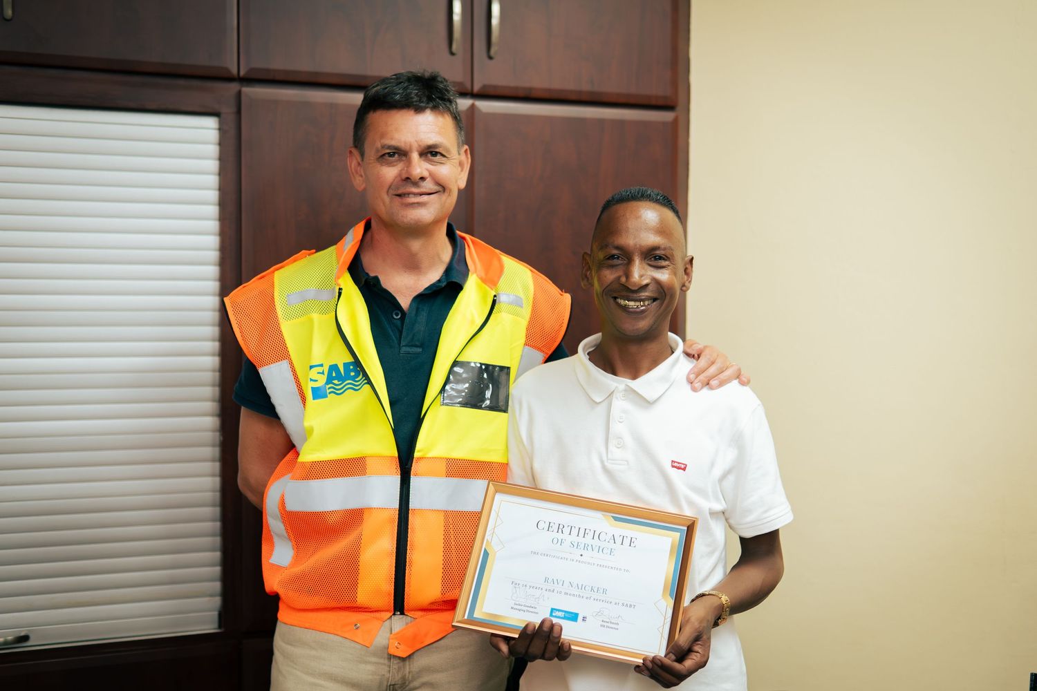 Two men pose together, one in an orange safety vest, the other holding a certificate of achievement indoors.