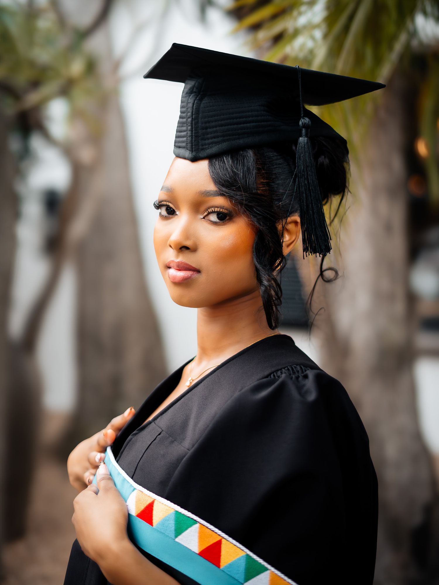 Young woman in black graduation cap and gown with colorful sash poses outdoors for graduation portrait.