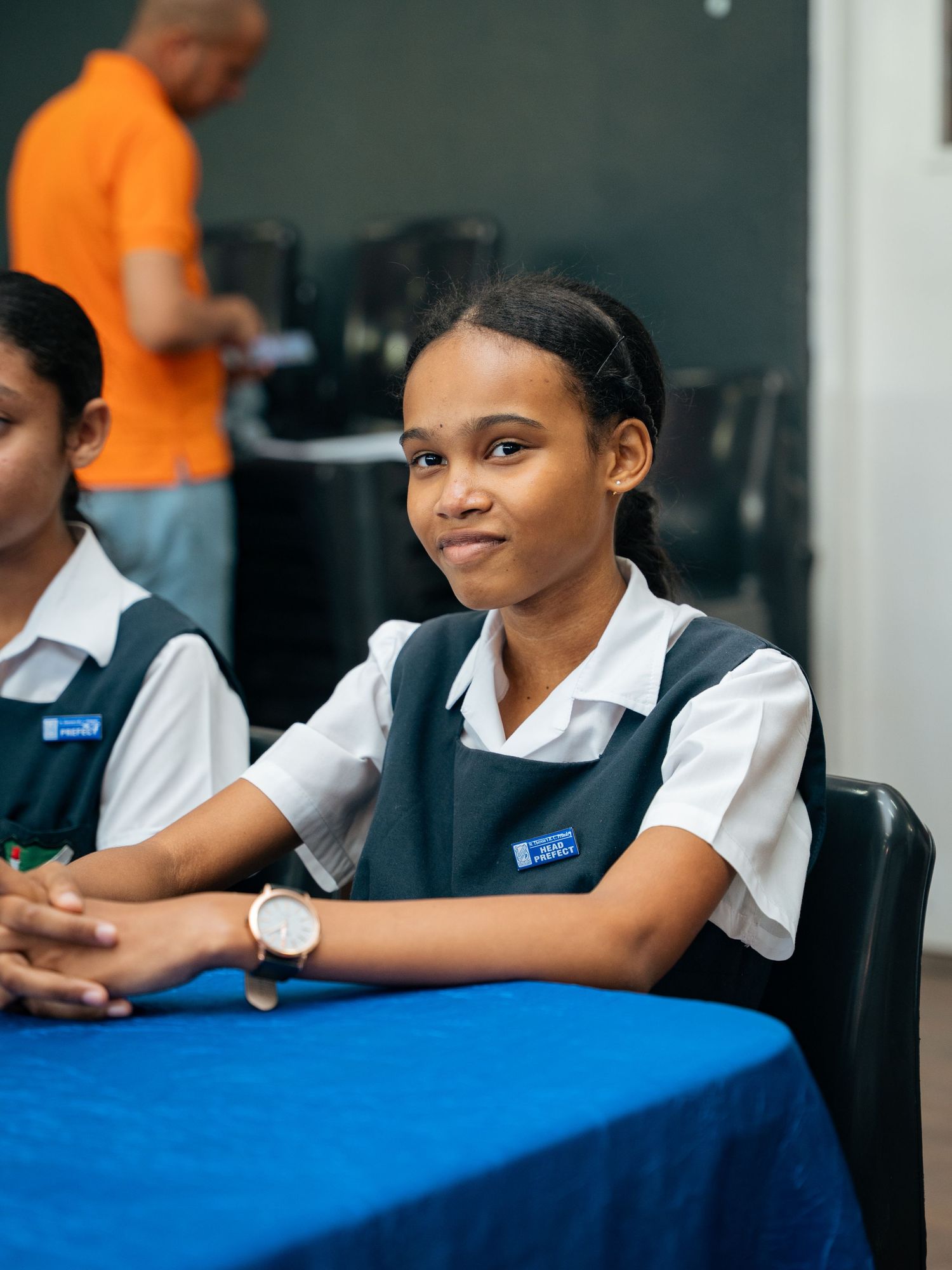 Young female student in school uniform smiles while seated at a blue table in a classroom setting.
