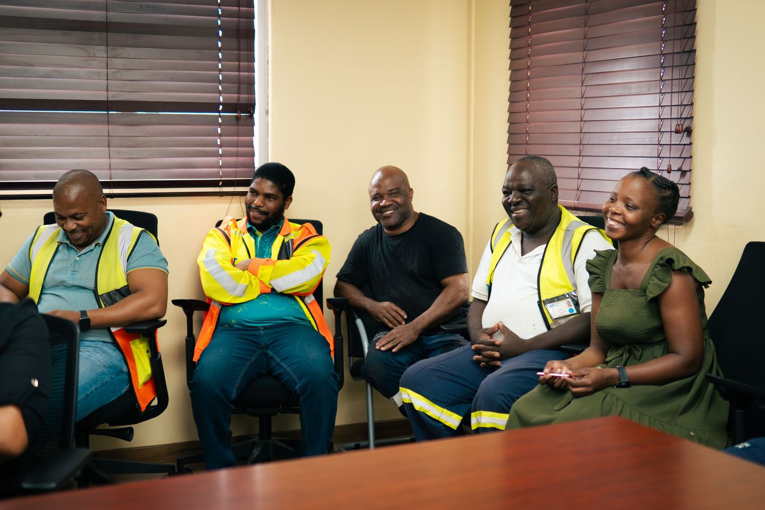 Group of five people sitting together laughing, some wearing safety vests, in an office setting.
