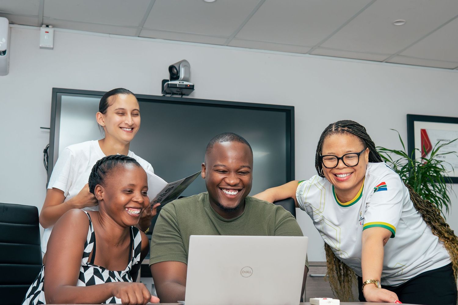 Four diverse colleagues smile while gathered around a Dell laptop in a modern office meeting room.