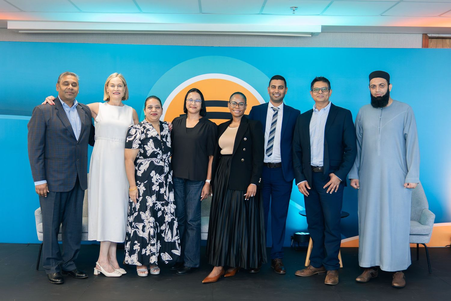 Group of eight diverse professionals posing together in front of a blue branded backdrop at a corporate event.