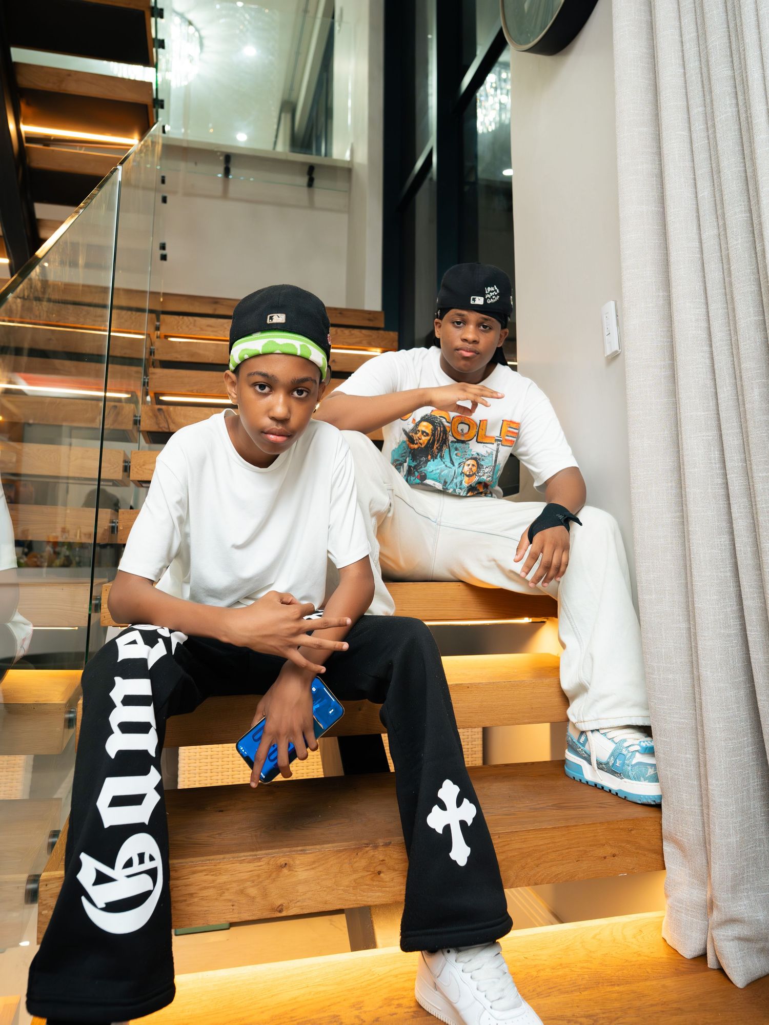 Two young men in streetwear sit on wooden stairs, wearing white tees, caps, and stylish sneakers in a modern interior.