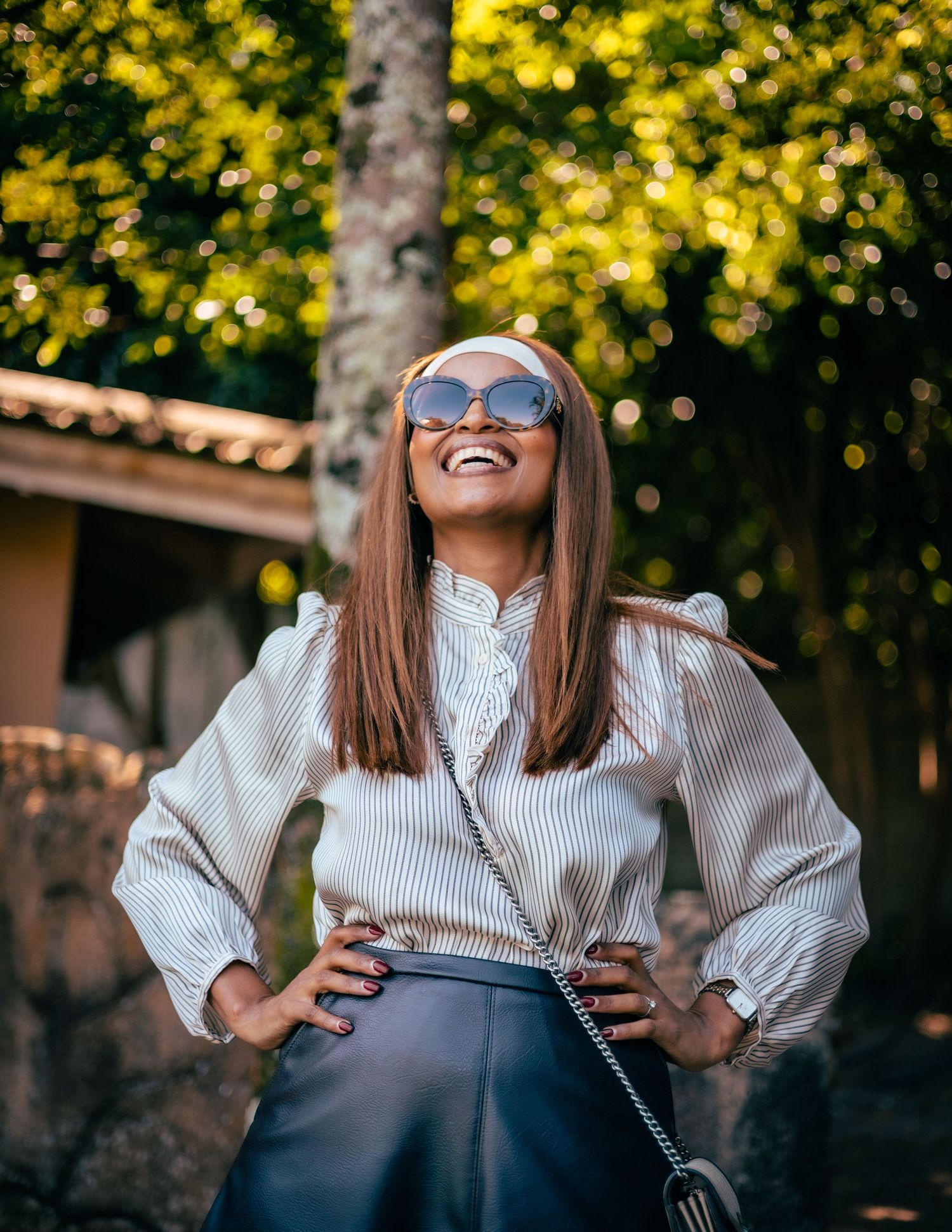 Smiling woman in striped blouse, leather skirt, and sunglasses posing outdoors with lush green trees in background.