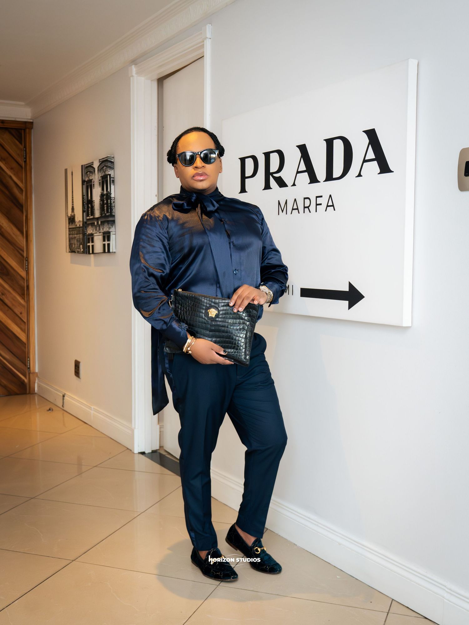 Stylish person posing by Prada Marfa sign in navy blue satin shirt, matching trousers, sunglasses, and black clutch bag.