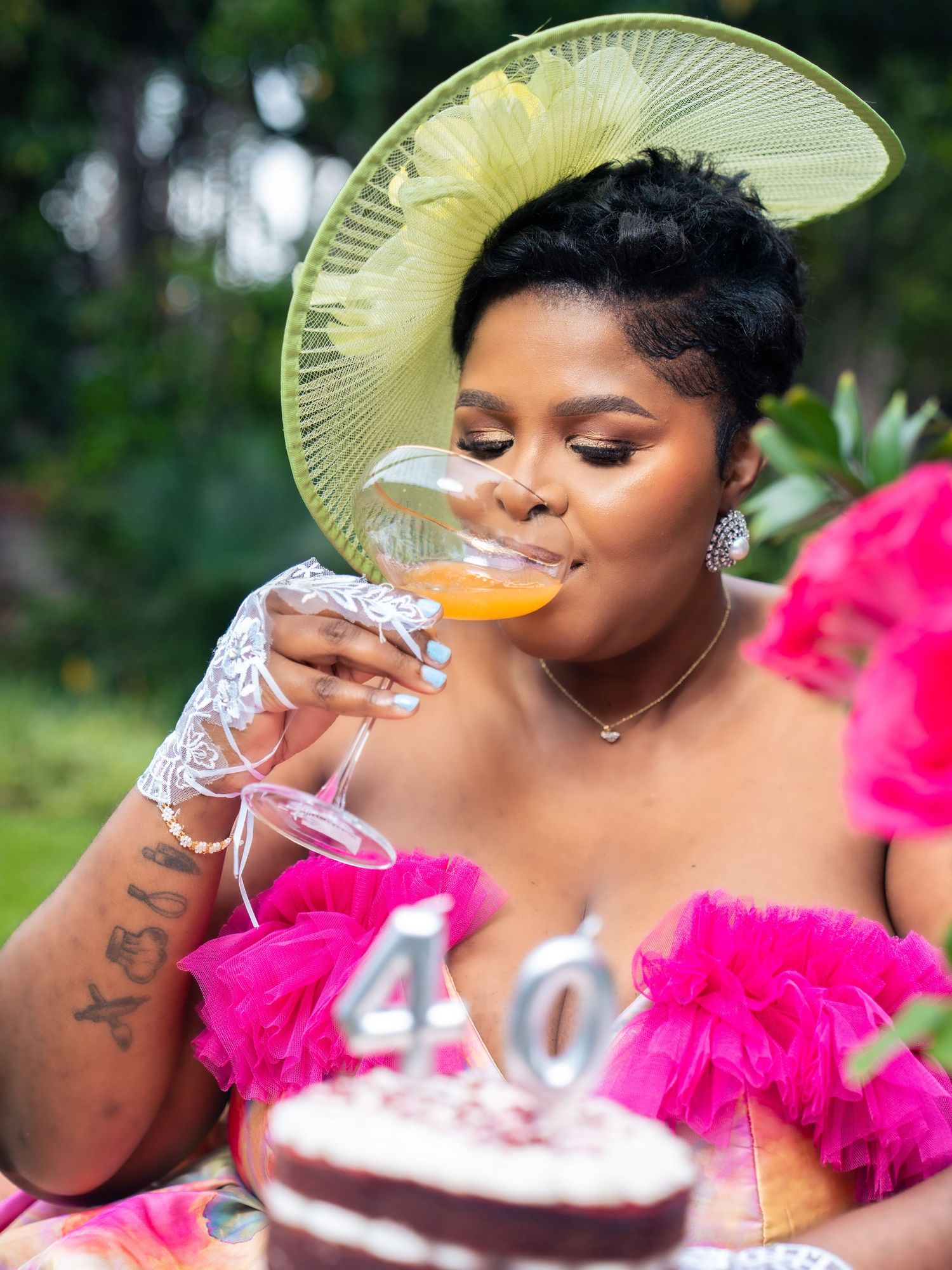 Woman in pink ruffled dress and yellow hat sips champagne at her 40th birthday celebration outdoors.